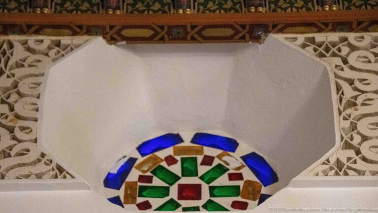Star-like medallion of blue, green, red, and amber glass framed by white carved plaster and painted wood.