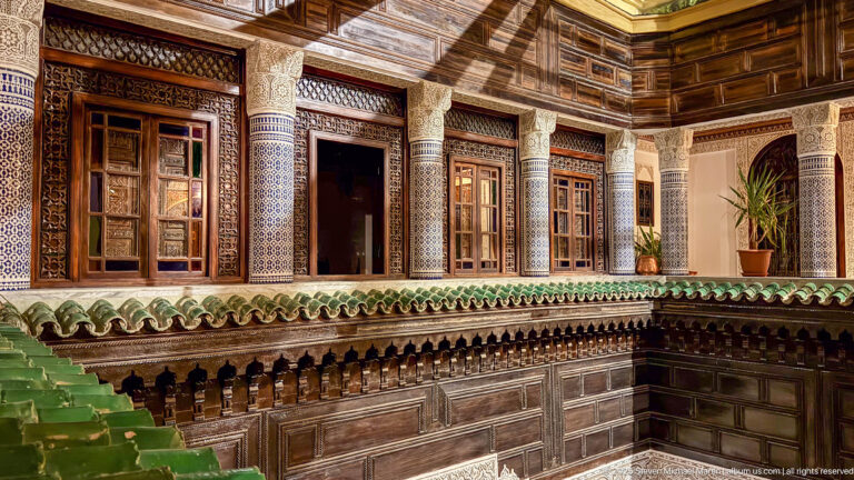 Upper riad gallery with dark carved wood, blue-and-white tiled columns, green-glazed eaves, and patterned windows.