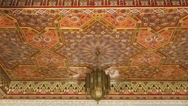 Upward view of a richly painted geometric wooden ceiling with octagonal panels, floral motifs, and a central hanging lantern.