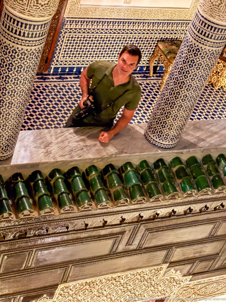 Man standing in the riad courtyard below tiled columns, mosaic walls, and green-glazed eaves, viewed from above.