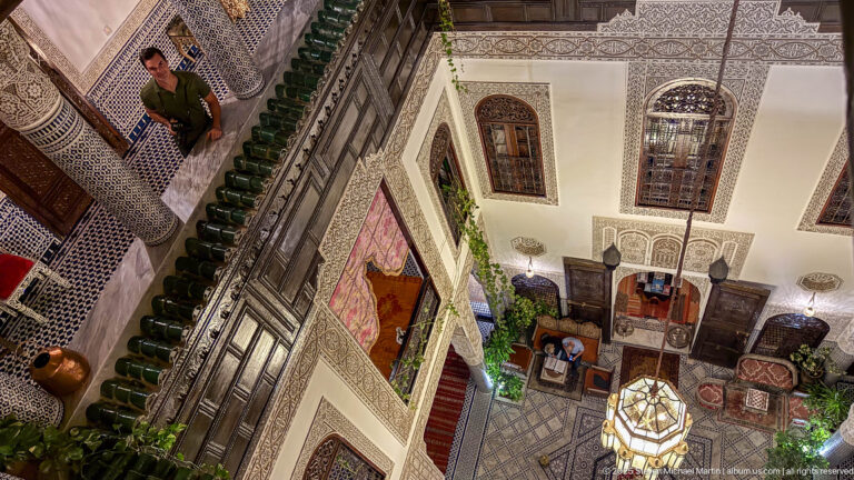 Steep downward view into the riad courtyard with hanging lantern, carved plaster, tiled columns, furnishings, and patterned floor.