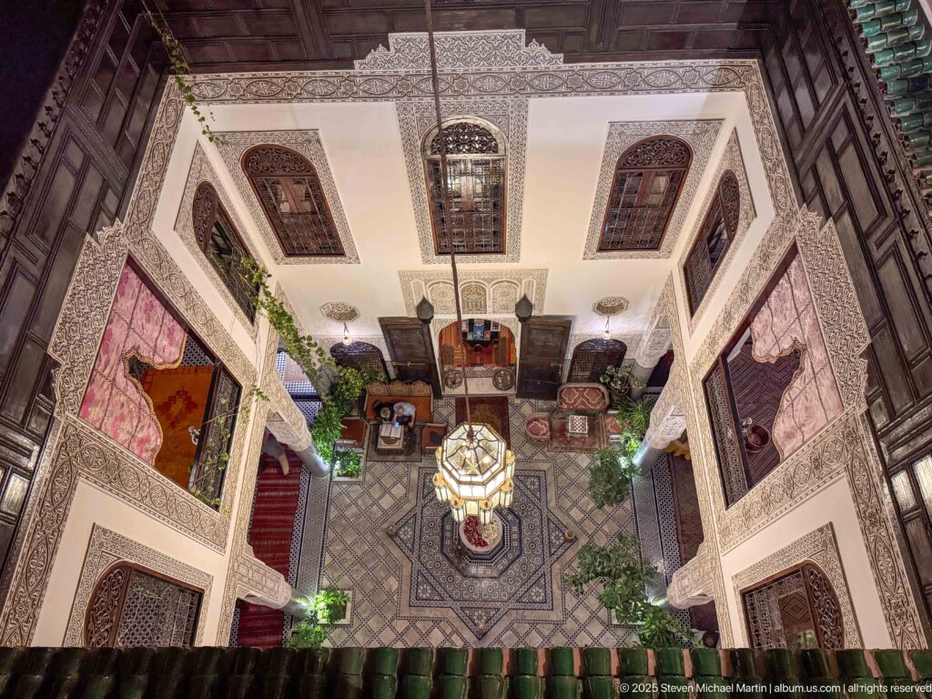 High-angle symmetrical view of the riad courtyard with central lantern, geometric floor, seating, plants, and carved wall openings.