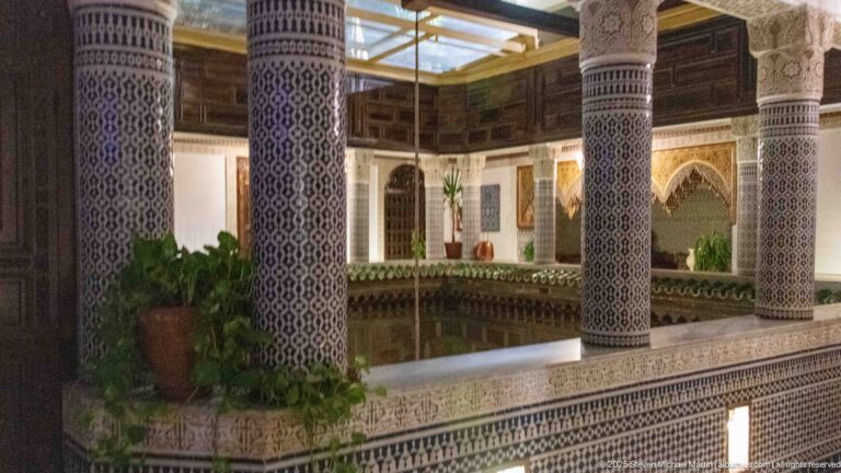 Upper riad gallery with blue-and-white tiled columns, dark timber walls, potted plants, and a glazed roof above.