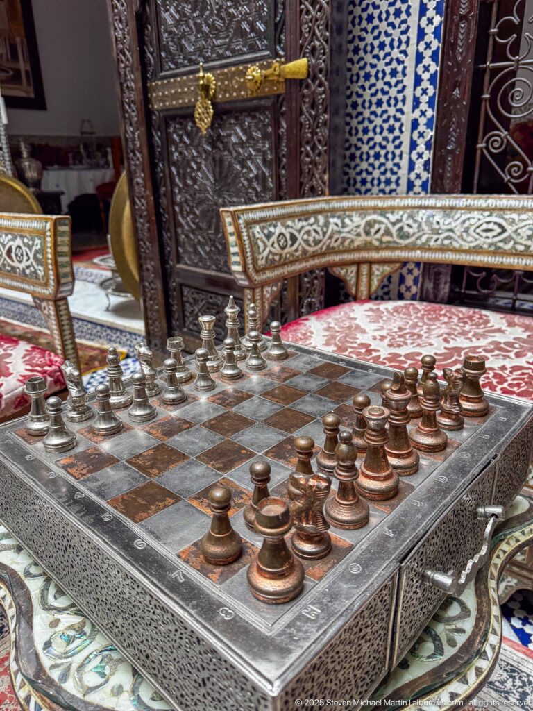 Decorative metal chessboard with turned chess pieces on an ornamented table amid patterned chairs and tiled walls.