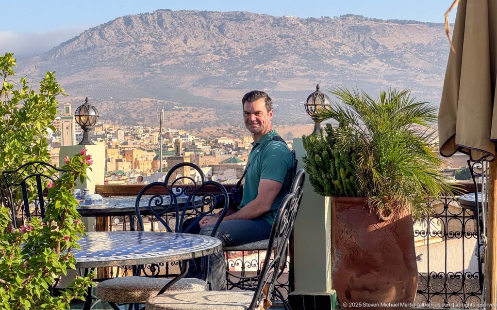 Seated man on a rooftop terrace with wrought-iron furniture and potted plants overlooking Fez and a rocky ridge.