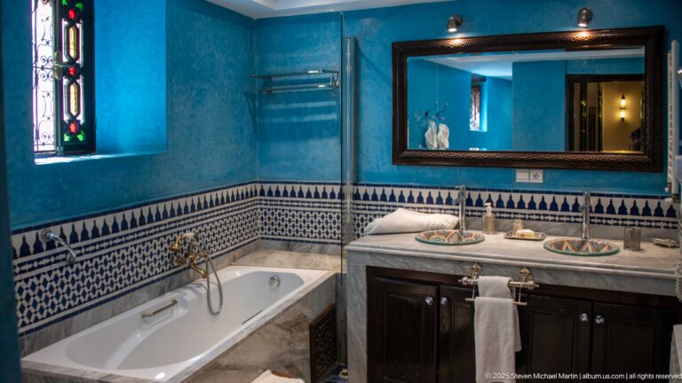 Blue guest bathroom with white bathtub, marble vanity, painted basins, framed mirror, and patterned zellij wall tile.