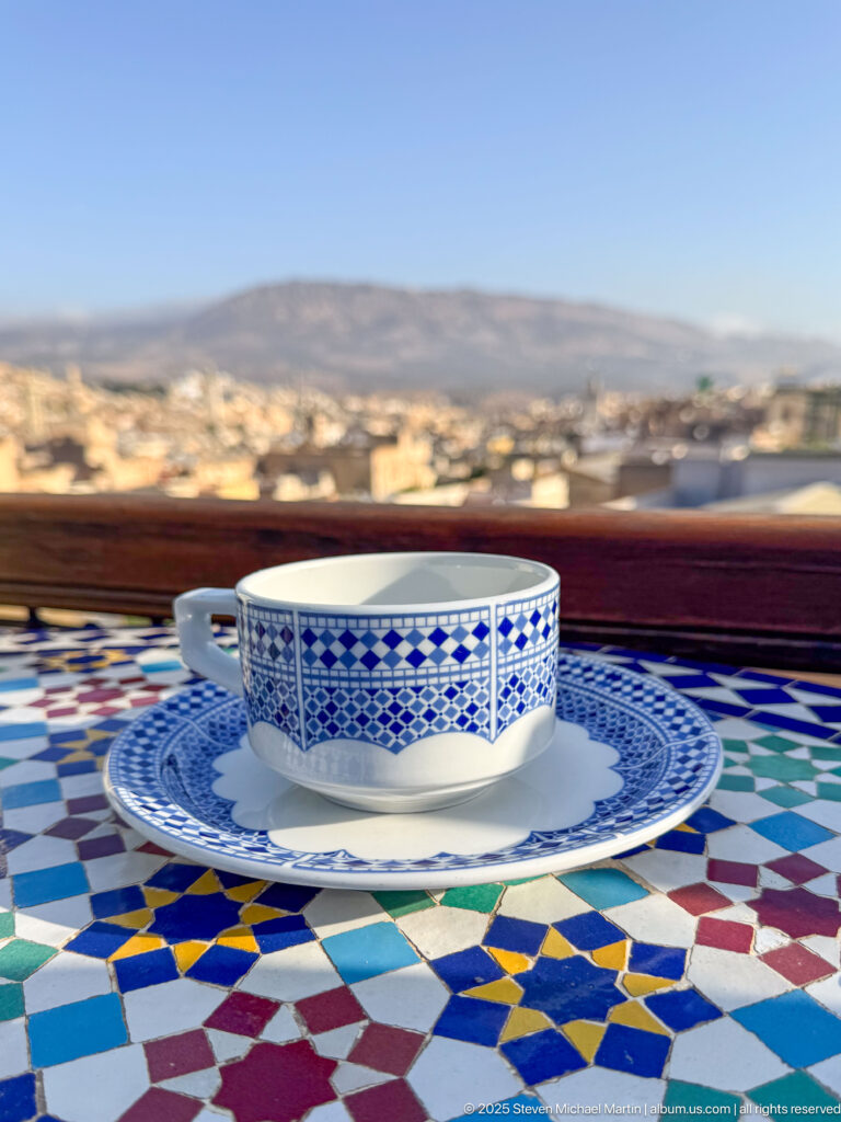 Blue-and-white ceramic cup and saucer on a multicolored geometric mosaic tabletop with a blurred cityscape beyond.