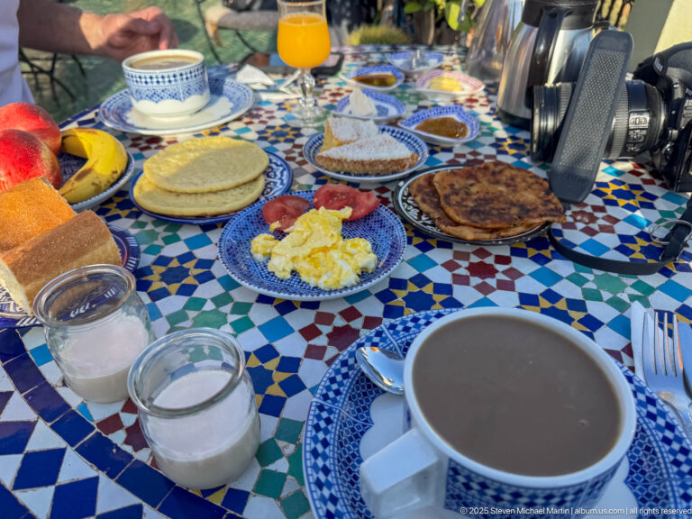 Breakfast spread with coffee, orange juice, eggs, flatbreads, pastries, fruit, yogurt, and blue-and-white dishes on a mosaic table.