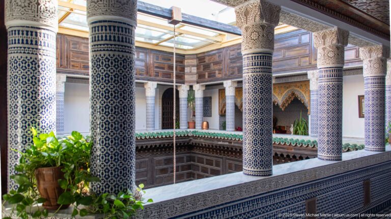 Upper riad gallery with blue-and-white tiled columns, carved plaster capitals, dark timber walls, green eaves, and skylight above.