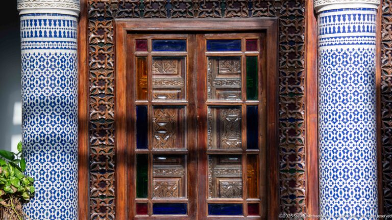 Carved wooden doorway with rectangular relief panels, colored-glass inserts, ornate wood frame, and blue-and-white zellij.