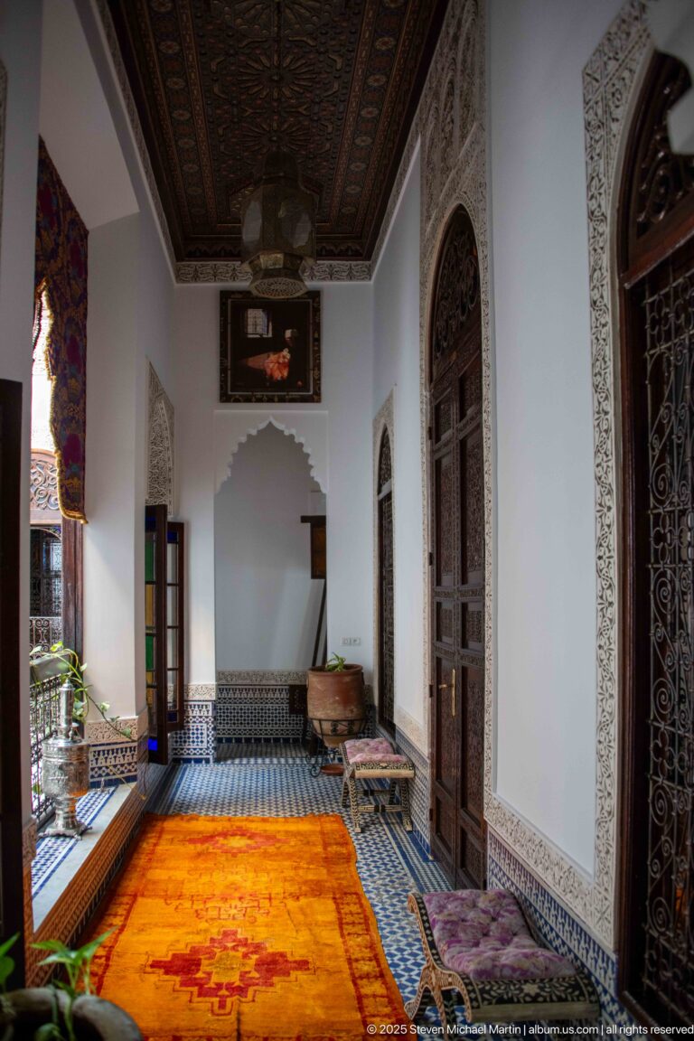 Long riad room with orange carpet, low side seating, blue-and-white tiled dado, carved wood doors, and decorated ceiling.