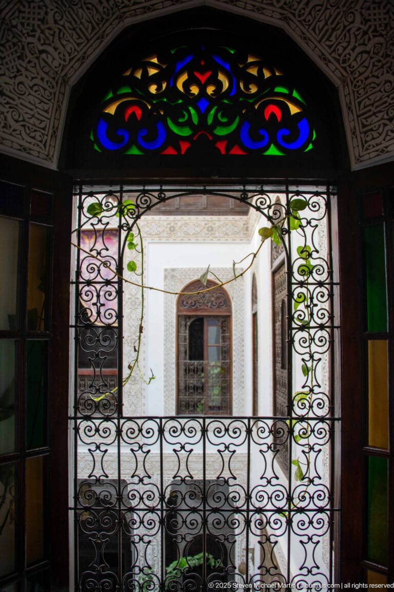 Scrolling wrought-iron grille beneath a semicircular multicolored glass transom, looking toward a bright interior courtyard.