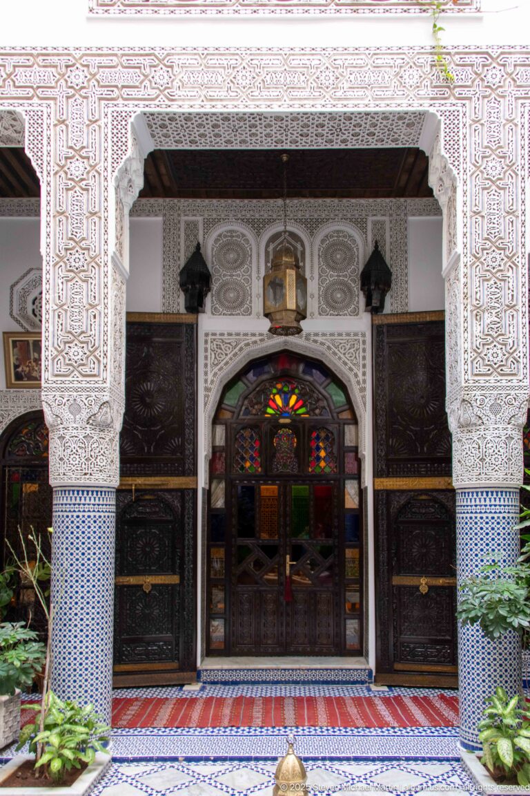 Symmetrical carved plaster portal framing a multicolored glazed doorway between dark wooden panels, tiled bases, lanterns, and plants.