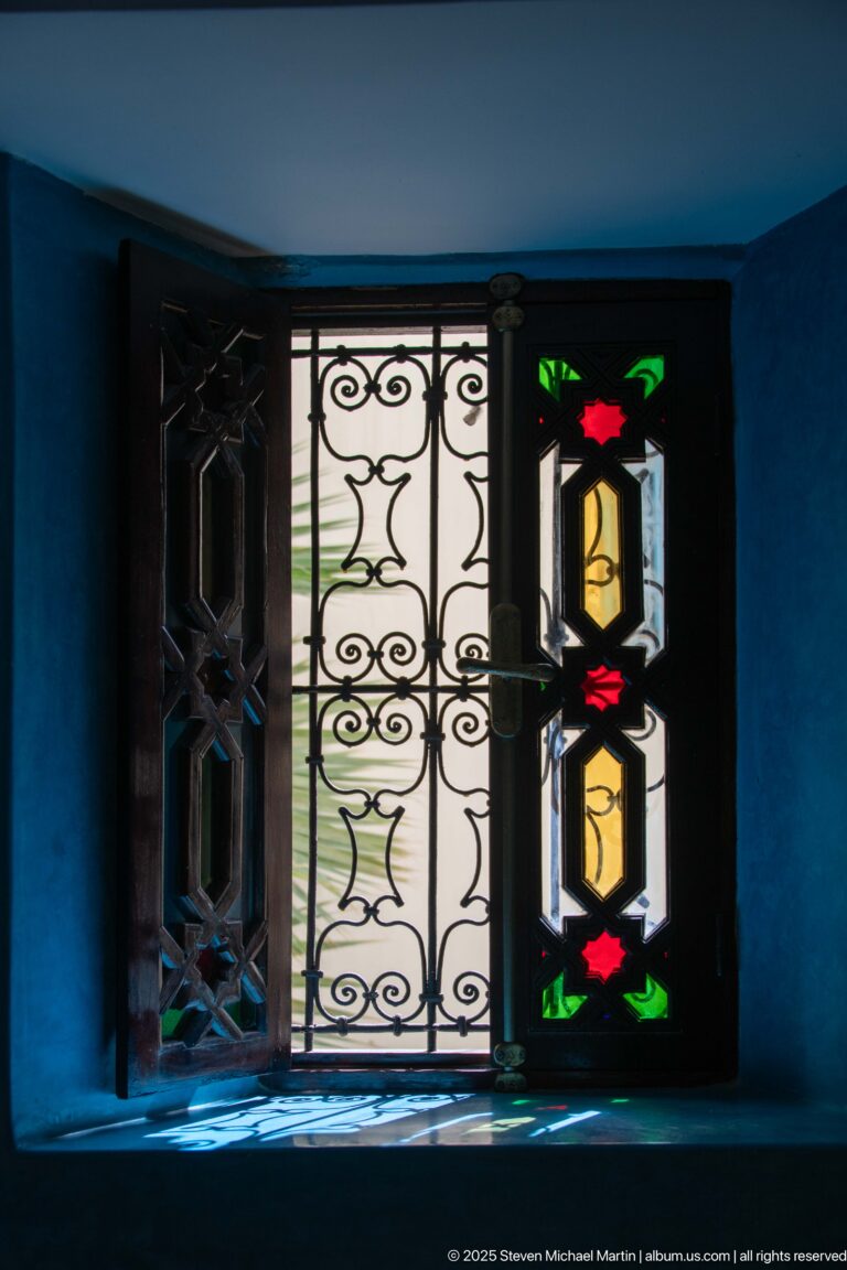 Small window with carved wood shutters, wrought-iron grille, and multicolored glass panels set in a vivid blue wall.