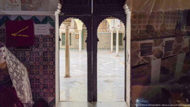 Dark exhibit room framing a bright courtyard doorway with a slender stone column and patterned tiled walls beyond.