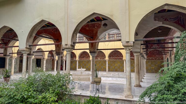 Pointed arches and slender columns overlooking a green palace courtyard with a small fountain in the distance.