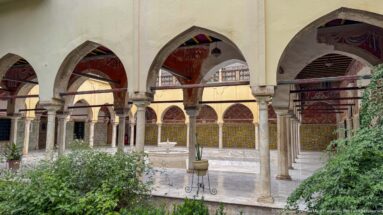 Pointed arches and slender columns overlooking a green palace courtyard with a small fountain in the distance.