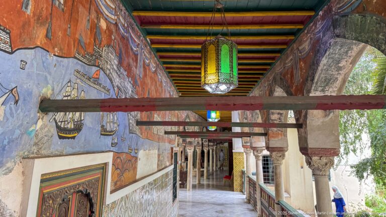 Long palace gallery with wall murals, tiled lower walls, hanging lanterns, columns, and a brightly striped painted ceiling.