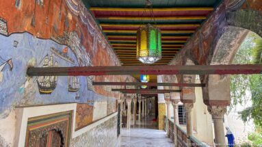 Long palace gallery with wall murals, tiled lower walls, hanging lanterns, columns, and a brightly striped painted ceiling.