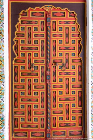 Red double wooden doors with interlocking yellow and black geometric bands, diamond panels, and a patterned tile border.