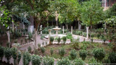 Leafy palace courtyard with a small central fountain, clipped shrubs, flowers, trees, and arcaded galleries behind.