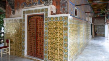 Oblique palace gallery wall with red geometric door, yellow-green tilework, weathered murals, and polished floor.
