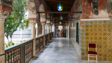 Long shaded gallery with stone columns, painted railing, yellow-green tile walls, ceiling beams, lanterns, and courtyard greenery.