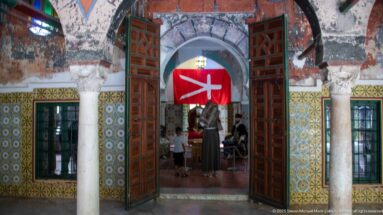 Open red-brown doors beneath an arch, flanked by carved columns and tilework, with visitors and a red hanging beyond.