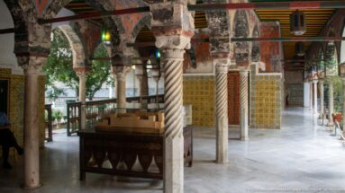 Palace arcade with mixed stone columns, spiral-fluted column, painted arches, tiled walls, timber ceiling, and display case.