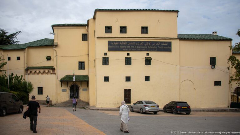 Exterior of Ahmed Bey Palace with pale walls, small windows, green-tiled eaves, museum sign, pedestrians, and parked cars.
