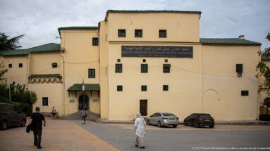 Exterior of Ahmed Bey Palace with pale walls, small windows, green-tiled eaves, museum sign, pedestrians, and parked cars.