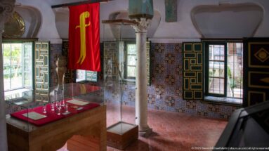 Decorated palace room with patterned tiles, green painted shutters, stone column, glass display case, and large red banner.