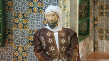 Male museum mannequin wearing a white turban and dark embroidered jacket against patterned tile walls.