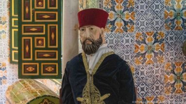 Male museum mannequin in a red fez and dark gold-trimmed garment beside geometric woodwork and floral tiles.