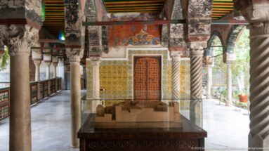 Architectural model in a display case beneath a palace arcade with stone columns, painted arches, tiles, and red door.