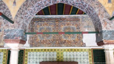 Marble arch and columns framing a weathered mural above yellow-green patterned tiles and painted ceiling beams.