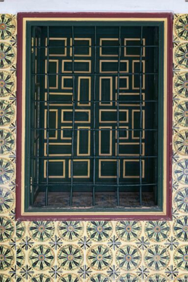 Dark geometric metal grille over a green-and-gold window, surrounded by repeating yellow-green patterned tiles.