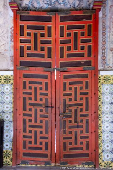 Red double wooden door with black interlocking geometric panels, narrow decorative borders, and patterned tilework.