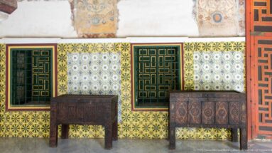 Two decorated wooden tables beneath green geometric window grilles against a wall of repeating floral ceramic tiles.