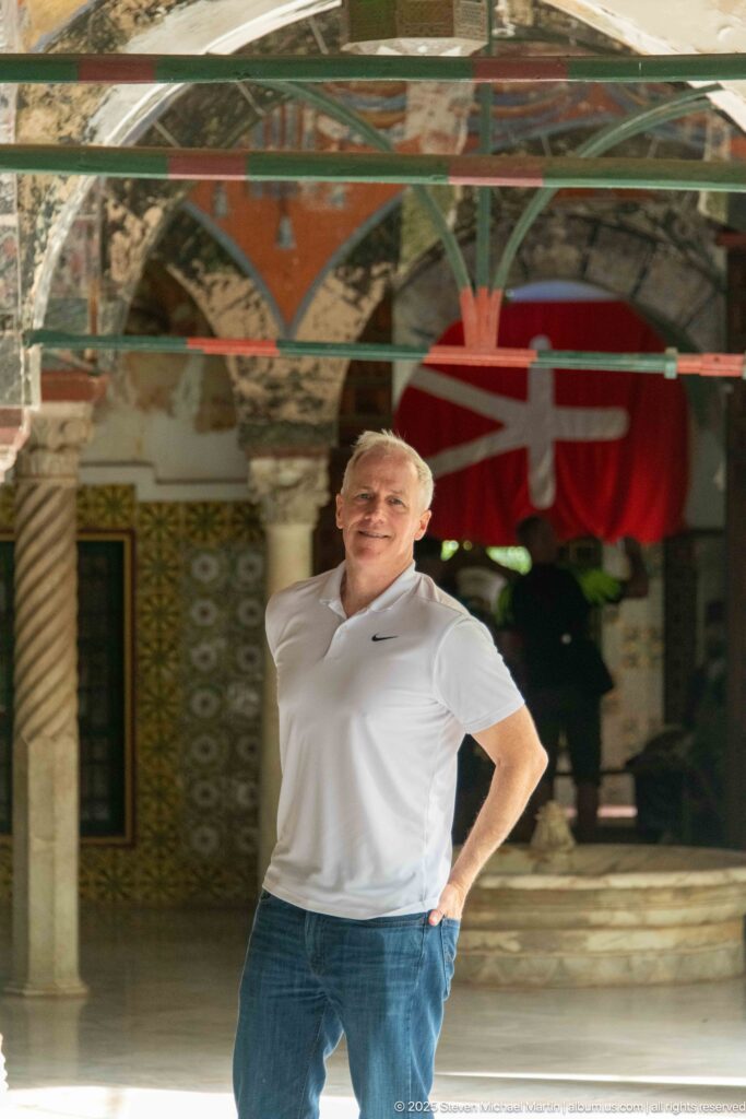 Man in a white polo shirt standing beneath a decorated palace arcade with columns, tilework, and a red hanging behind.
