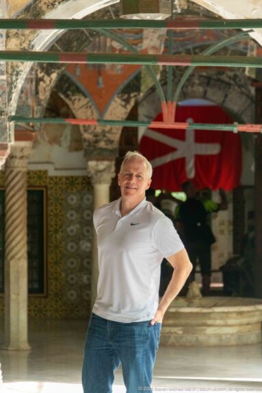 Man in a white polo shirt standing beneath a decorated palace arcade with columns, tilework, and a red hanging behind.
