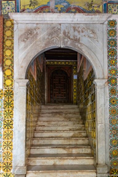 Marble steps rising through a rounded stone arch, with colorful tiled walls and a dark wooden doorway at the top.