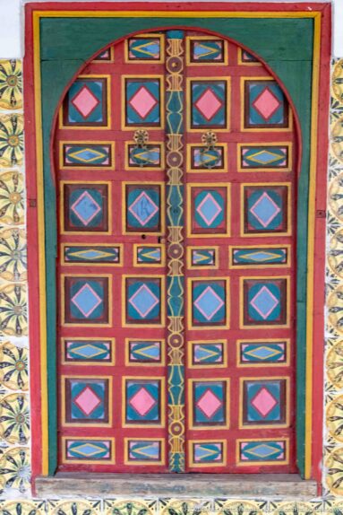 Arched double wooden door painted red, green, yellow, and blue with rows of geometric panels and diamond motifs.