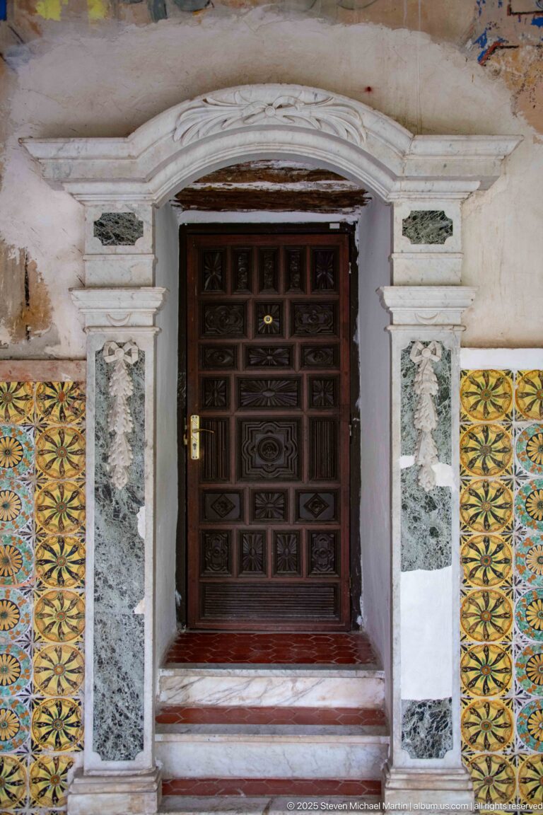 Dark paneled wooden door framed by pale carved stone, narrow gray stone inserts, and yellow-green patterned tilework.