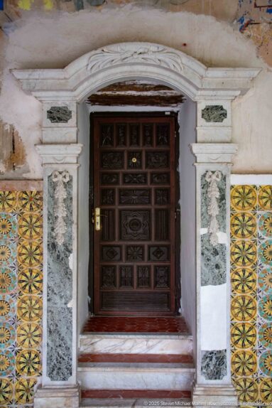 Dark paneled wooden door framed by pale carved stone, narrow gray stone inserts, and yellow-green patterned tilework.