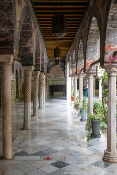 Long palace gallery with pointed arches, stone columns, striped painted ceiling, marble floor, and potted plants beside the courtyard.
