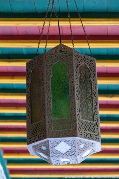 Polygonal hanging lantern with pierced metal panels and colored glass suspended by chains beneath a striped painted ceiling.