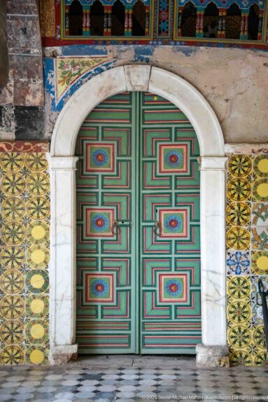 Green double door with red geometric bands and painted medallions beneath a rounded stone arch, surrounded by patterned tiles.