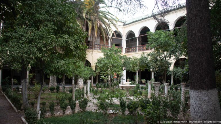 Leafy palace courtyard with small fountain, mature trees, palms, and two-story arcaded galleries surrounding the garden.