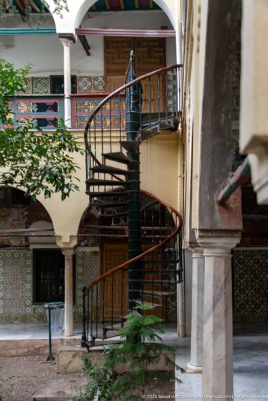 Dark metal spiral staircase in a palace courtyard surrounded by tiled walls, painted doors, columns, galleries, and foliage.